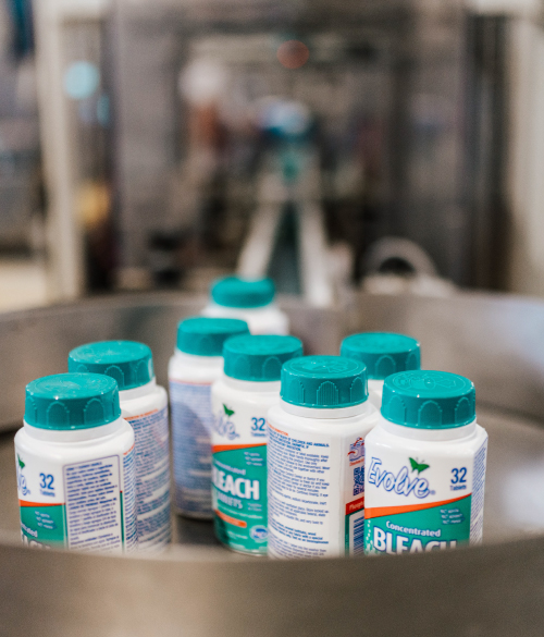 Varios envases de pastillas desinfectantes de la marca Evolve, alineados en una bandeja metálica dentro de una instalación industrial. Los frascos tienen tapas verde agua y etiquetas en tonos verde y blanco. Se puede leer 'Evolve', '32' y 'Concentrated BLEACH TABLETS', lo que indica que contienen 32 tabletas de lejía concentrada. El fondo está desenfocado, destacando el enfoque en los productos.